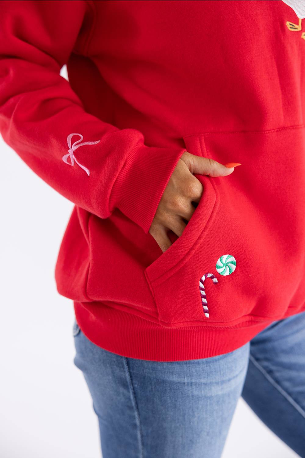 A close-up of a person wearing a bright red hoodie with a white embroidered bow on the sleeve and a candy cane patch on the pocket, paired with blue jeans.
