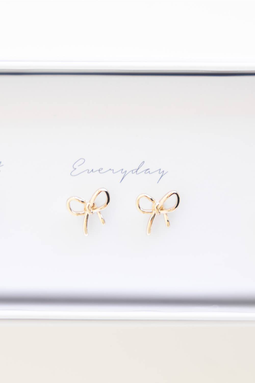 A pair of small gold bow stud earrings displayed on a white jewelry box lid with the word 'Everyday' written in cursive.
