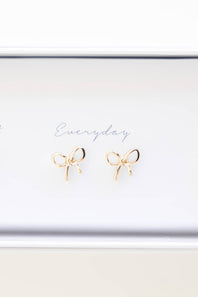 A pair of small gold bow stud earrings displayed on a white jewelry box lid with the word 'Everyday' written in cursive.