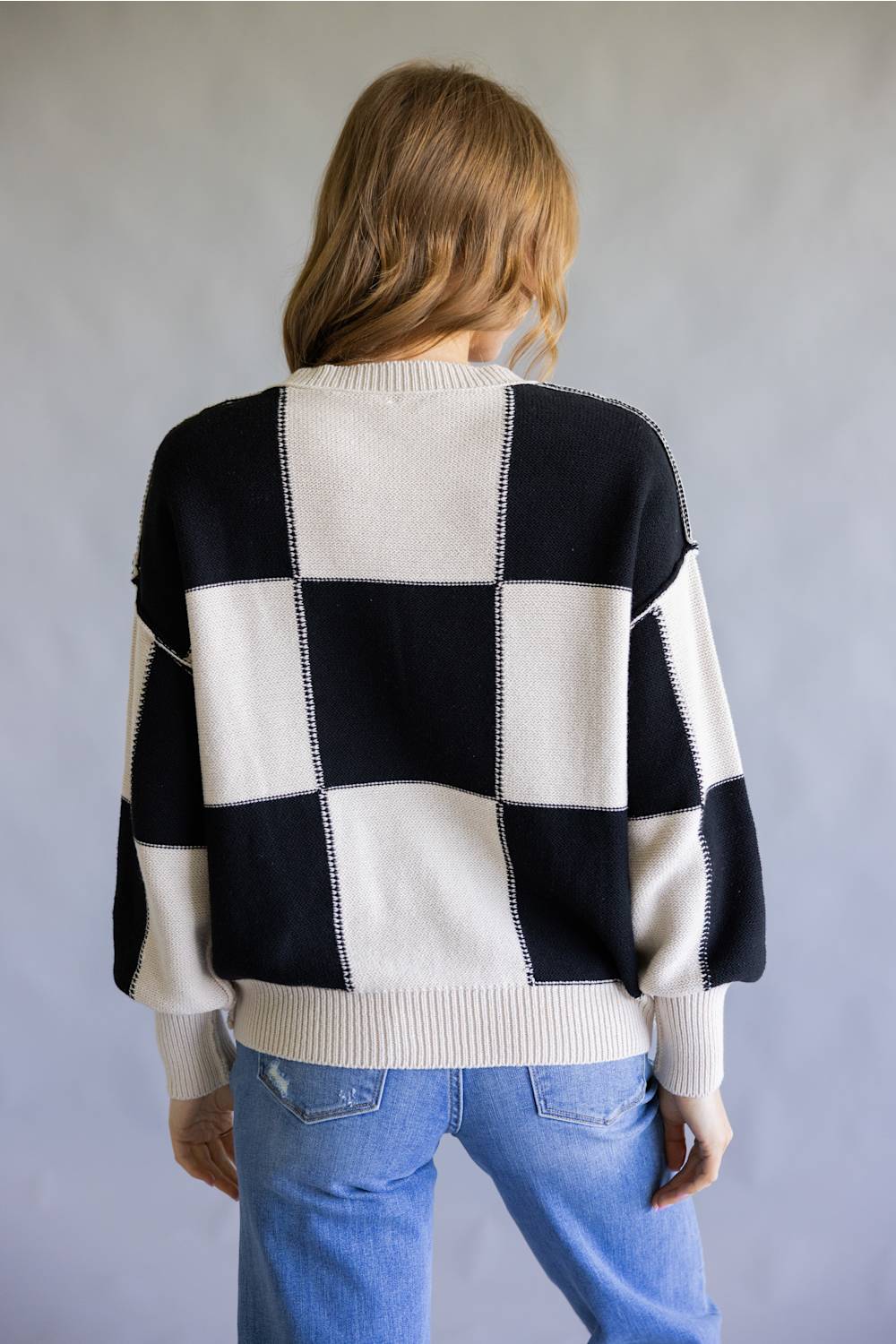 A woman viewed from behind wearing a black and off-white checkered knit sweater and light blue jeans, standing against a neutral gray background.