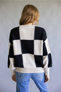 A woman viewed from behind wearing a black and off-white checkered knit sweater and light blue jeans, standing against a neutral gray background.