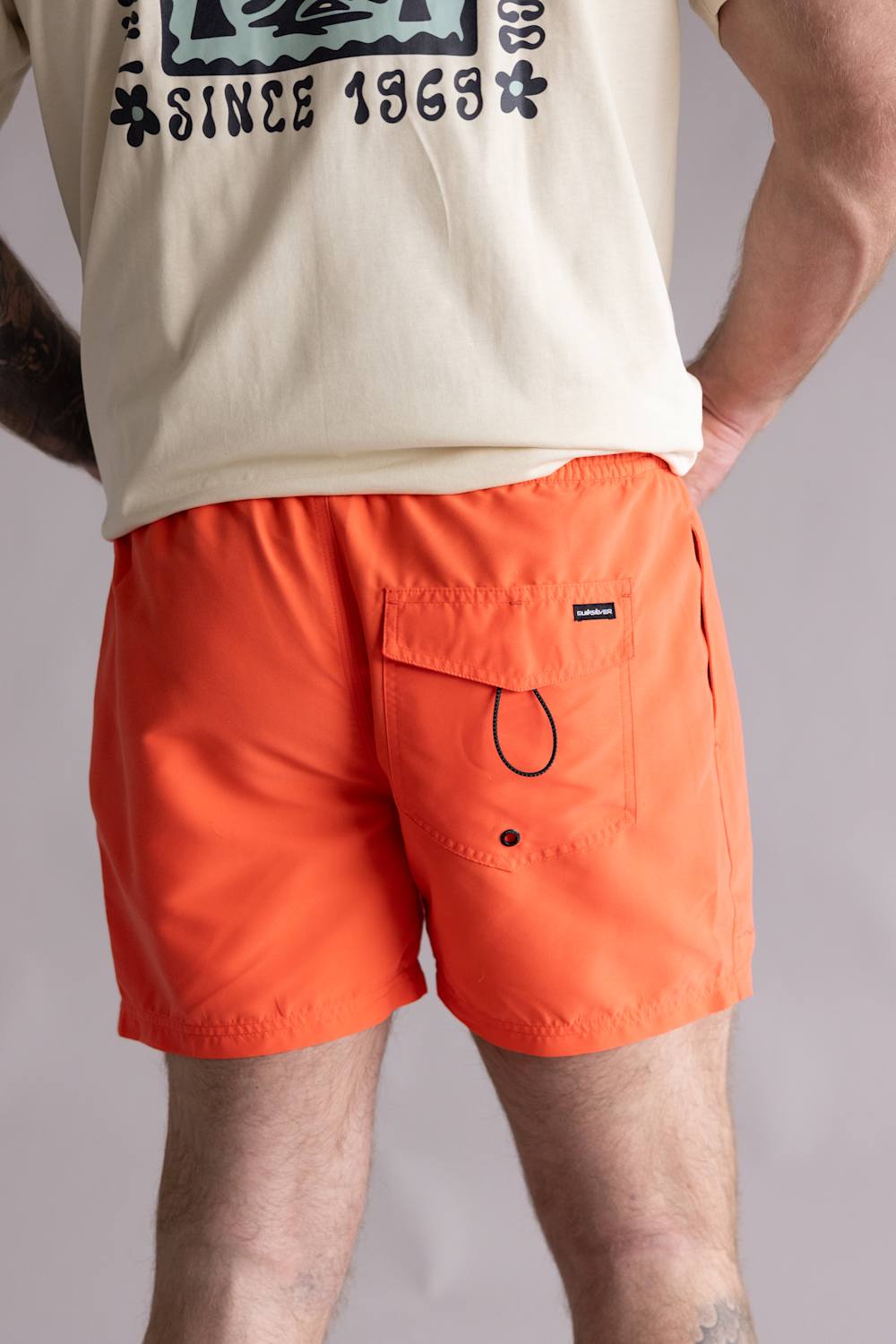 Back view of a person wearing a beige t-shirt with a graphic design and orange shorts on a plain background. The shorts are the Quiksilver Everyday Solid Volley 15" Swim Shorts for Men in Hot Coral