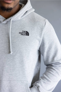 A close-up of a person wearing a light gray heather pullover hoodie with a black embroidered 'The North Face' logo on the left chest, set against a neutral gray background.
