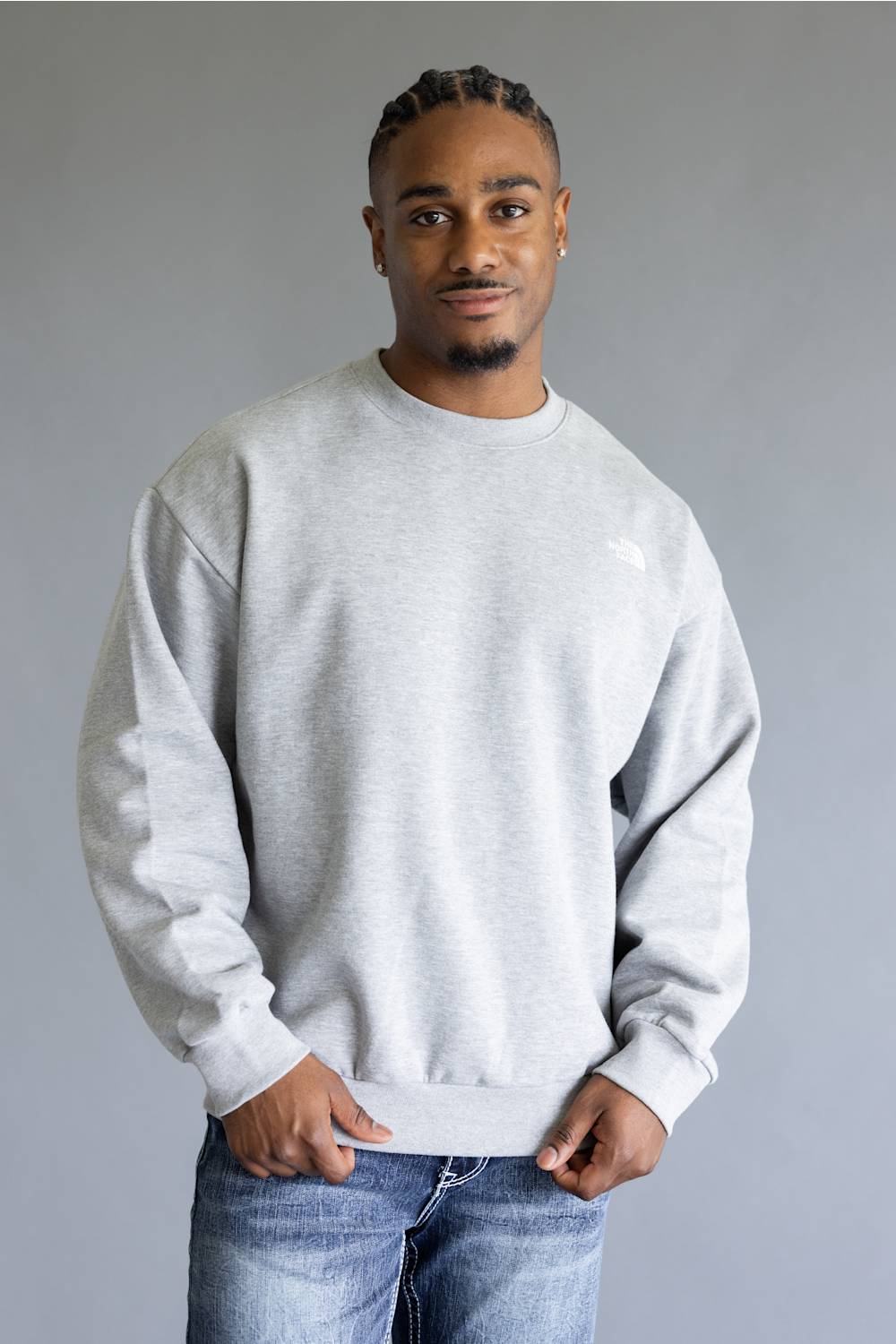 A man wearing a light gray crewneck sweatshirt and blue jeans, standing against a neutral gray background.