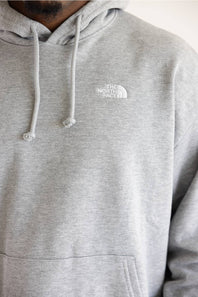 A close-up of a person wearing a light heather gray pullover hoodie with white drawstrings and a small white embroidered The North Face logo on the left chest.