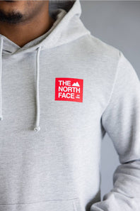 A man wearing a light gray heather pullover hoodie with a red 'The North Face' logo patch on the left chest, paired with blue jeans, against a solid gray studio background.