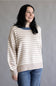 A woman wearing a beige and white horizontally striped knit sweater with a contrasting blue crew neck, paired with blue jeans, against a plain gray background.
