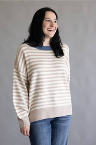 A woman wearing a beige and white horizontally striped knit sweater with a contrasting blue crew neck, paired with blue jeans, against a plain gray background.