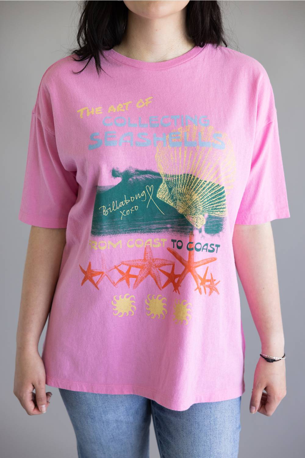A person wearing a pink graphic T-shirt with a vintage-style shell and wave print, paired with blue jeans, against a plain gray background.