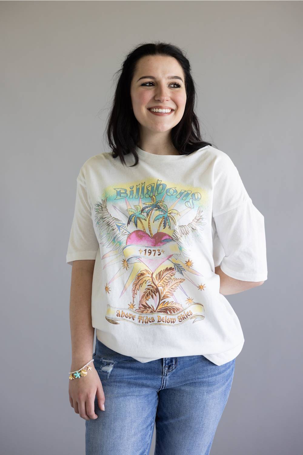 A woman wearing a vintage-style white graphic t-shirt with a colorful tropical design and blue jeans, standing against a plain gray background.