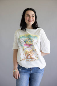 A woman wearing a vintage-style white graphic t-shirt with a colorful tropical design and blue jeans, standing against a plain gray background.