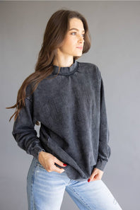 A woman wearing a dark gray, acid-washed long-sleeve mock neck sweatshirt and light blue jeans, posing against a solid gray studio background.