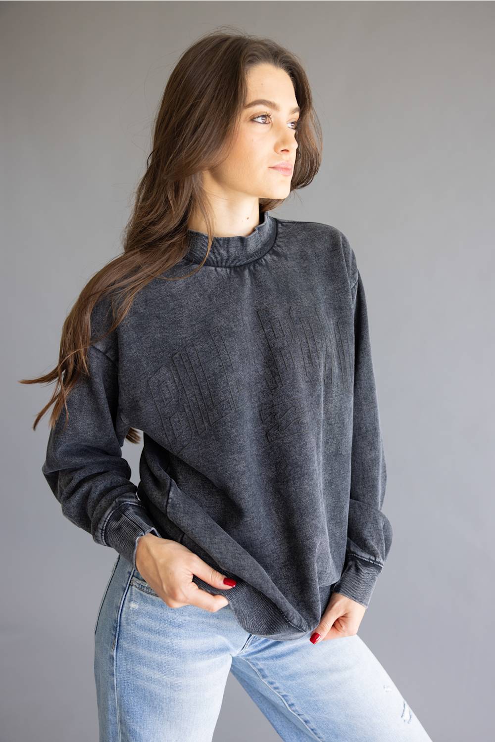 A woman wearing a dark gray, acid-washed long-sleeve mock neck sweatshirt and light blue jeans, posing against a solid gray studio background.