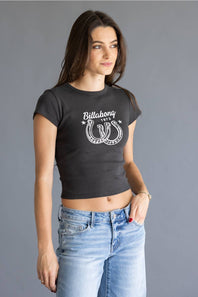 A woman wearing a black short-sleeve crop top with a white Billabong horseshoe graphic, paired with light blue jeans, against a solid gray studio background.