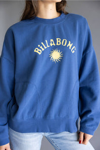 A person wearing a blue crewneck sweatshirt with 'Billabong' and a sun graphic printed in yellow on the chest, paired with light blue jeans against a gray studio background.