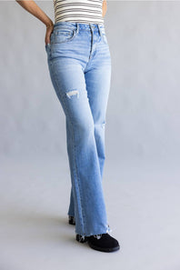A person wearing light-wash blue flare jeans with a distressed knee patch, paired with a striped top and black shoes, photographed against a plain light gray studio background.
