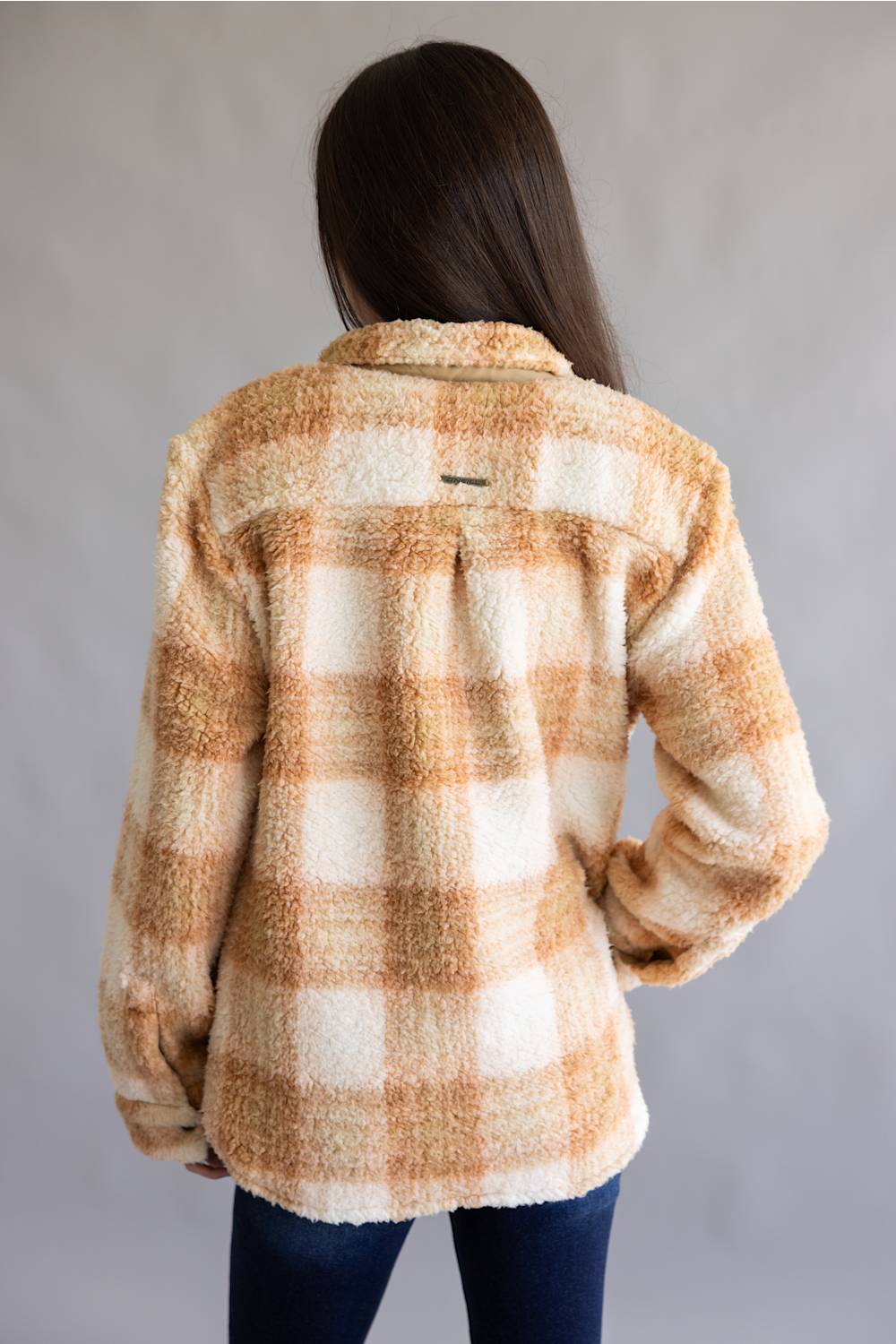 A person shown from the back wearing a fuzzy plaid flannel-style jacket in warm beige and cream tones, paired with dark blue jeans, against a plain light gray studio background.
