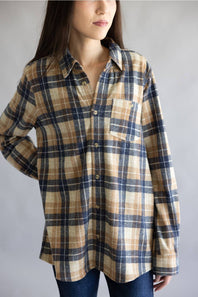 A woman wearing a long-sleeve button-up plaid shirt in beige, navy, and brown tones, paired with dark blue jeans, photographed against a neutral gray background.
