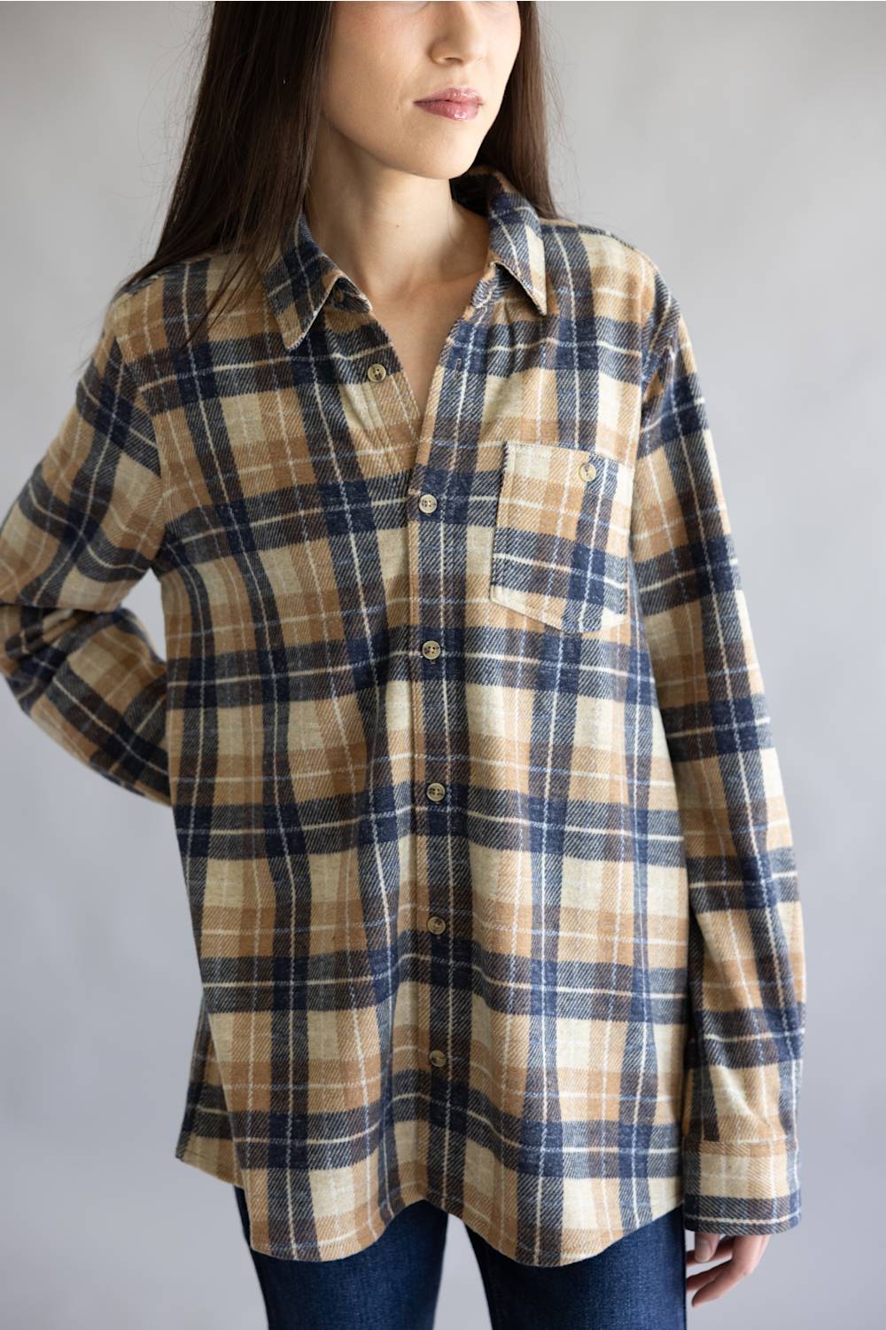 A woman wearing a long-sleeve button-up plaid shirt in beige, navy, and brown tones, paired with dark blue jeans, photographed against a neutral gray background.