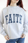 A woman wearing a light heather gray crewneck sweatshirt with the word 'FAITH' printed across the chest in navy blue block letters filled with white polka dots, paired with blue jeans.