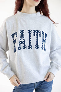 A woman wearing a light heather gray crewneck sweatshirt with the word 'FAITH' printed across the chest in navy blue block letters filled with white polka dots, paired with blue jeans.