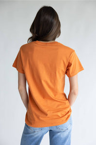 A person viewed from behind wearing a plain orange short-sleeve T-shirt and light blue jeans against a light gray studio background.