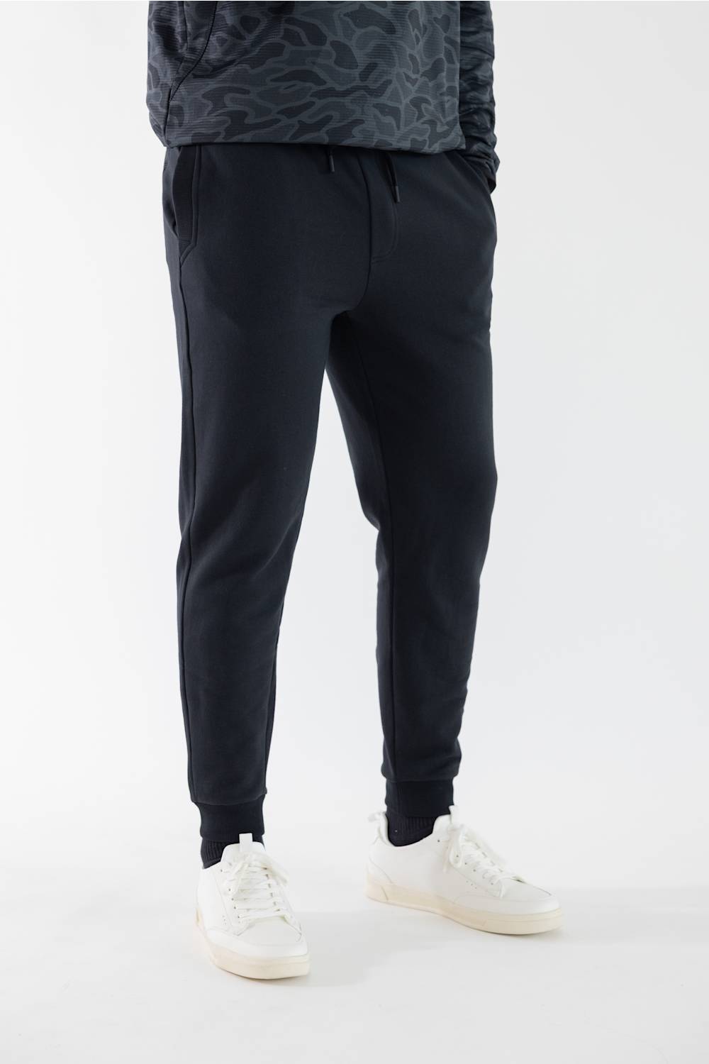 A person wearing  jogger-style pants with elastic cuffs, paired with white sneakers and a dark patterned top, shown against a plain light gray studio background.