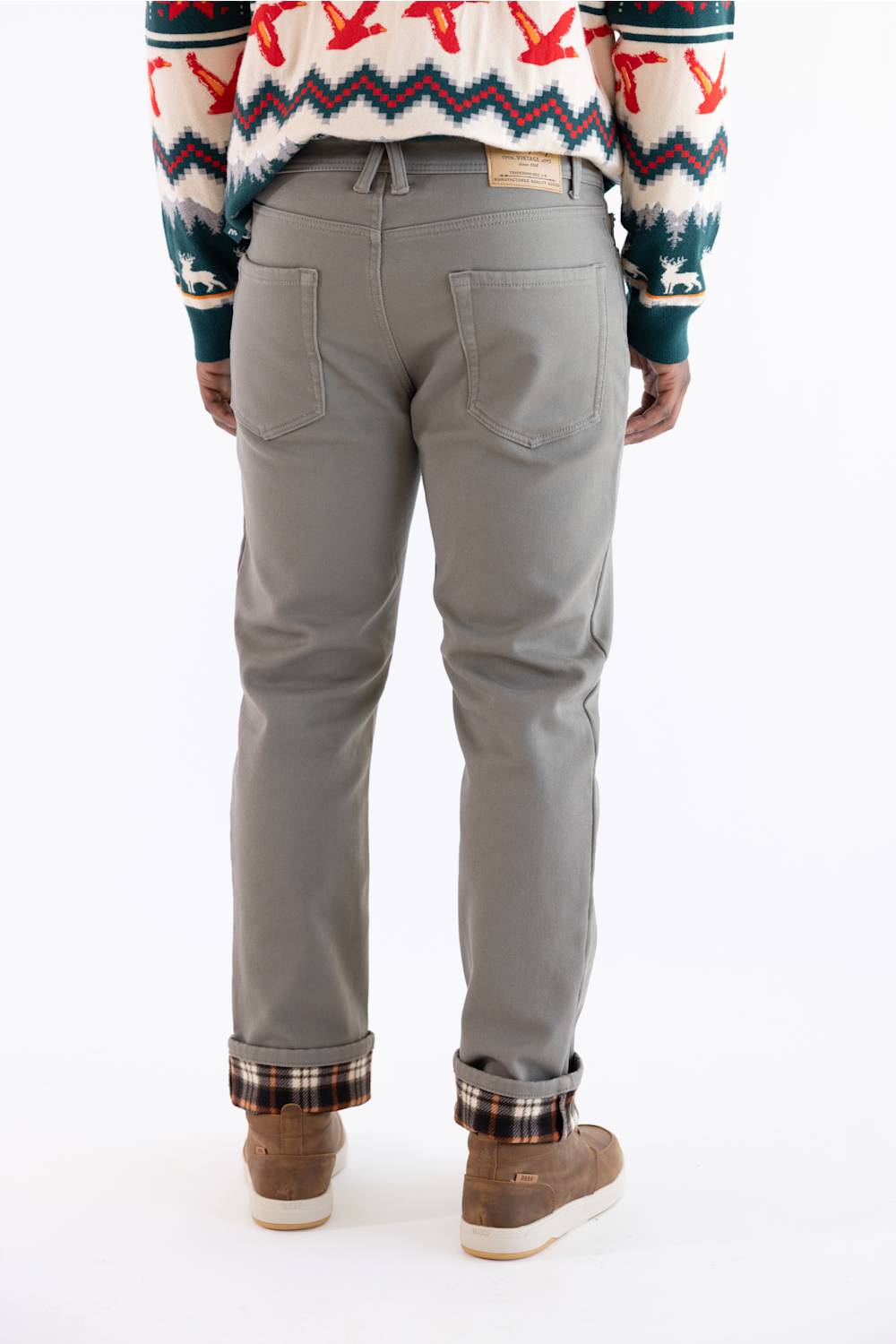 A person shown from the back wearing light gray slim-fit jeans with a patterned knit sweater and brown suede boots, photographed against a plain white studio background.