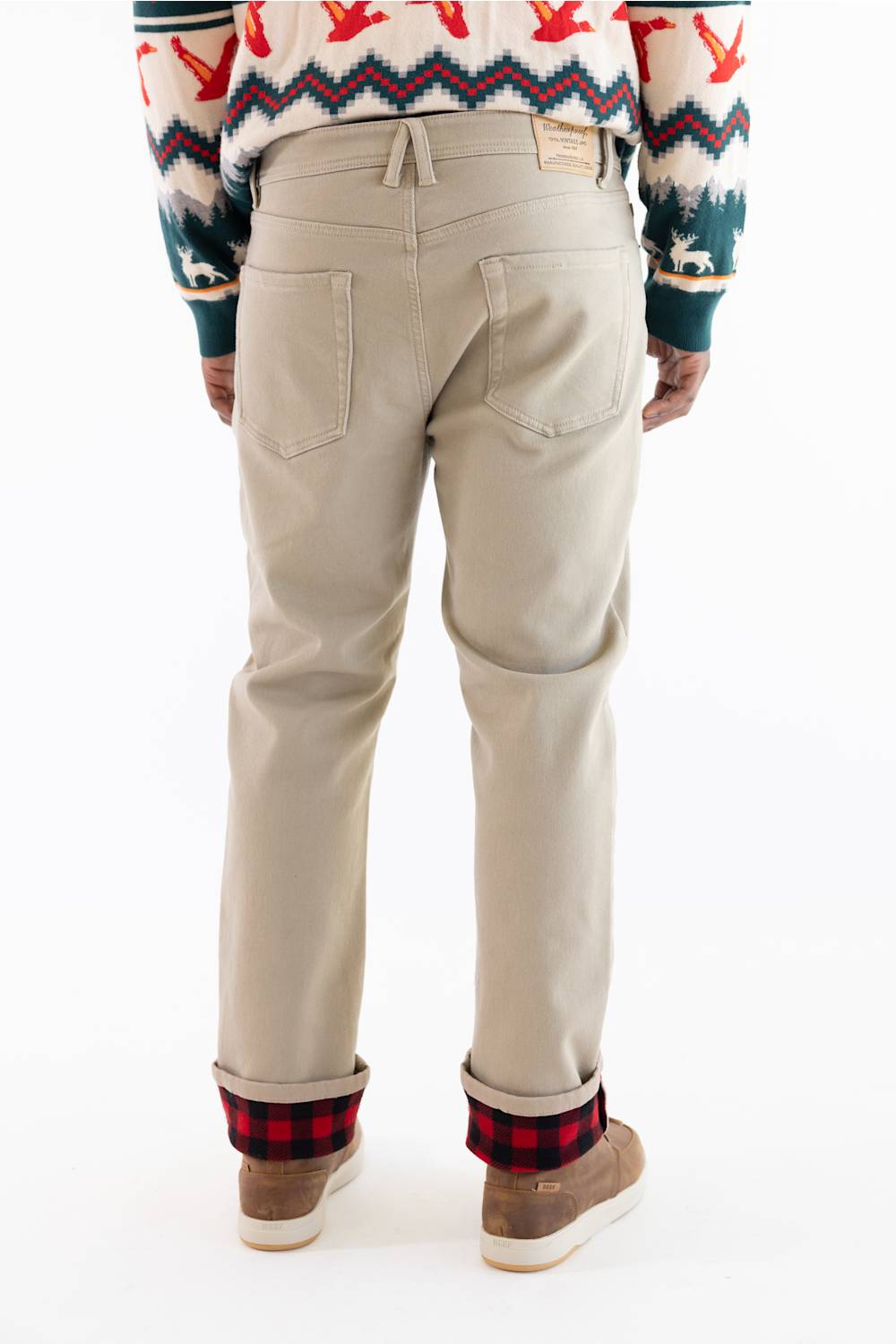Back view of a person wearing beige chino pants with a red and black buffalo plaid cuff, paired with a patterned sweater and brown shoes, on a white background.