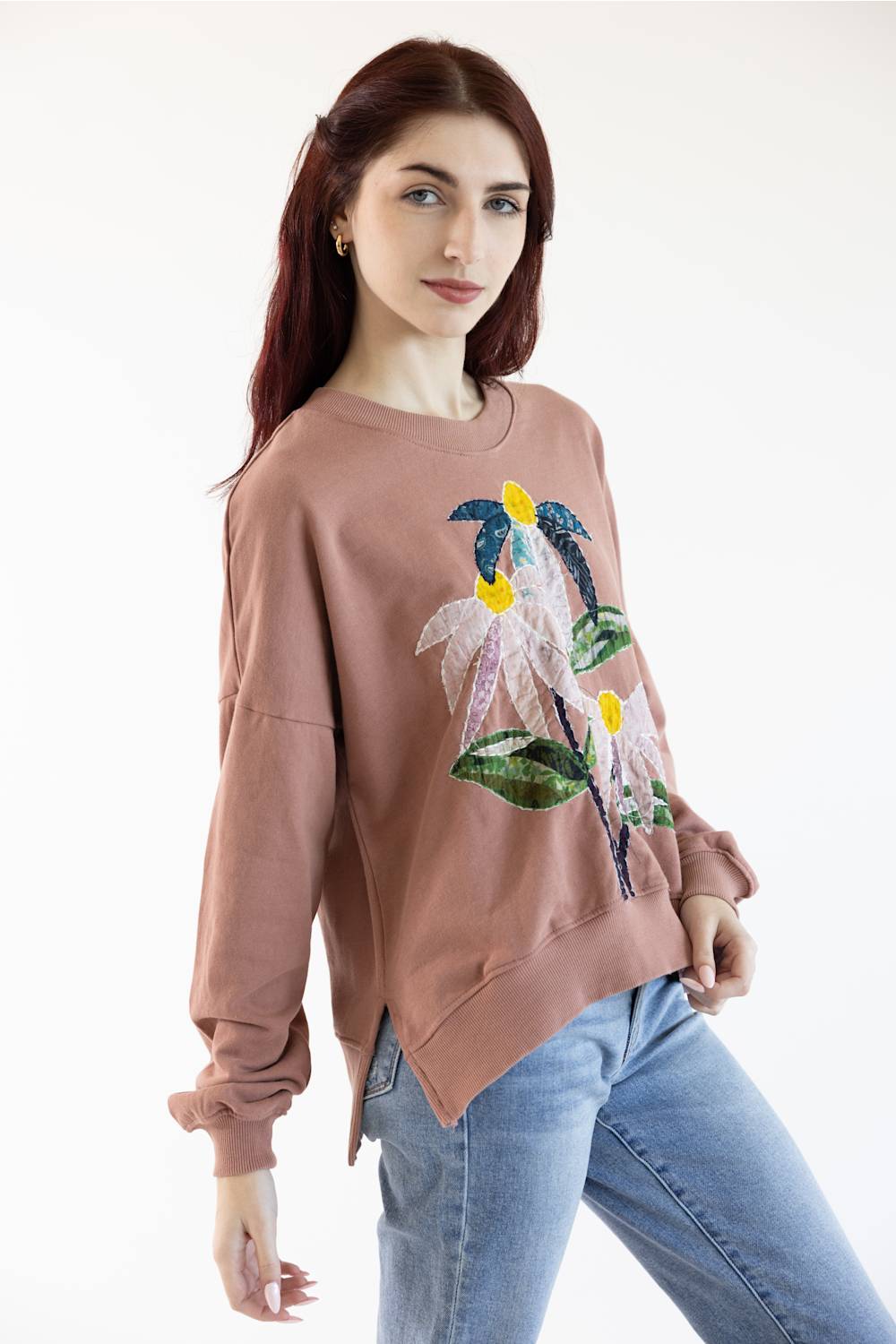 A woman modeling a dusty rose long-sleeve sweatshirt with a colorful floral sequin and bead applique on the front, paired with light blue jeans against a plain white background.
