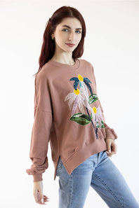 A woman modeling a dusty rose long-sleeve sweatshirt with a colorful floral sequin and bead applique on the front, paired with light blue jeans against a plain white background.