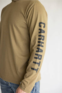 A close-up of a person wearing a tan long-sleeve shirt with 'CARHARTT' printed vertically in dark navy on the right sleeve, paired with blue jeans against a plain light background.