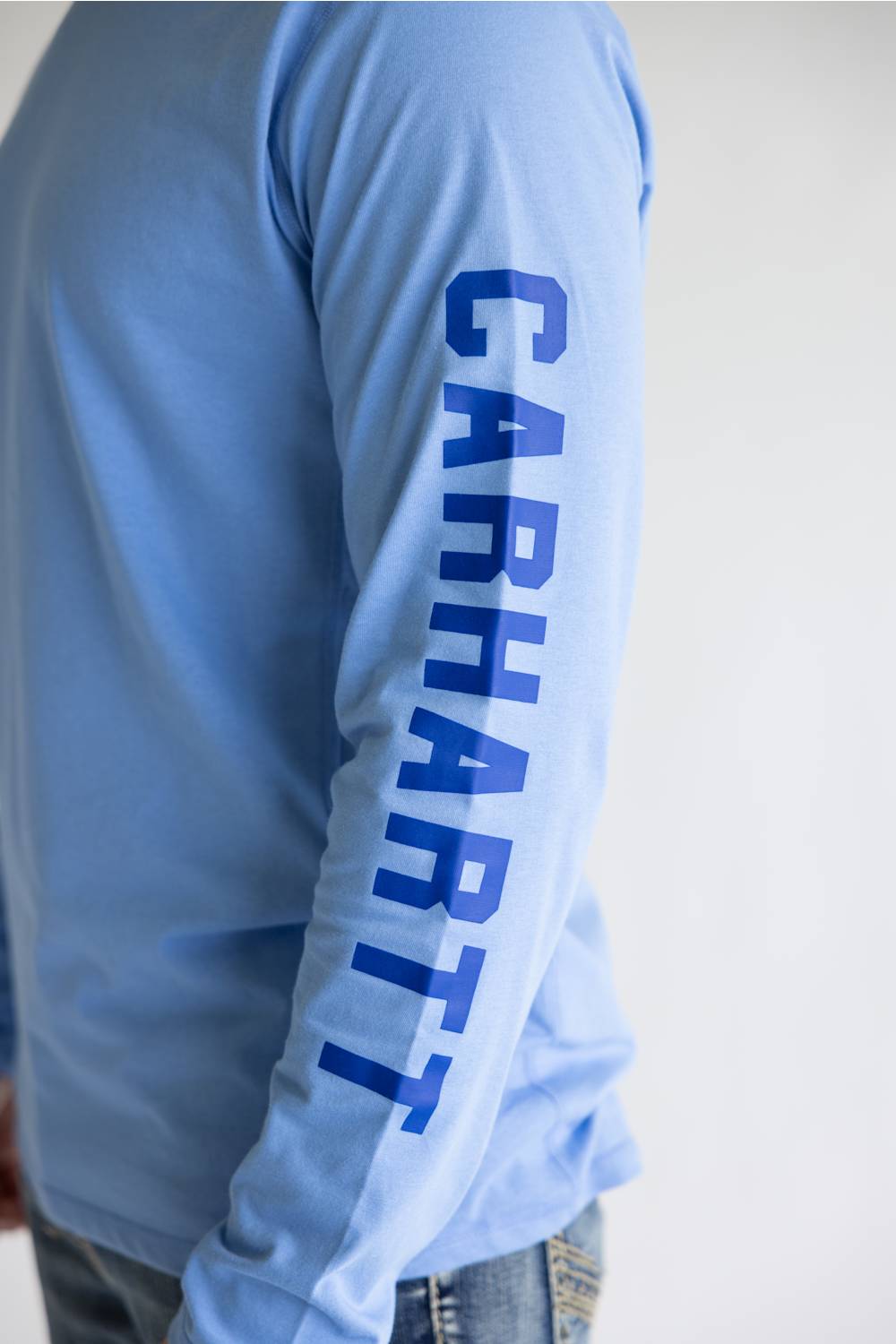 A close-up of a person wearing a light blue long-sleeve shirt with the brand name 'CARHART' printed vertically in dark blue down one sleeve, paired with blue jeans against a plain light background.