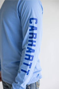 A close-up of a person wearing a light blue long-sleeve shirt with the brand name 'CARHART' printed vertically in dark blue down one sleeve, paired with blue jeans against a plain light background.