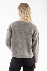 A woman viewed from behind wearing a heathered gray knit sweater and black pants against a plain white background.