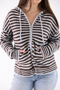 A woman wearing a brown and white striped zip-up hoodie with white drawstrings and light blue jeans, shown from the neck down against a plain white background.