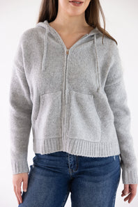 A woman wearing a light gray knit zip-up hoodie with drawstrings and front pockets, paired with blue jeans, against a plain white background.