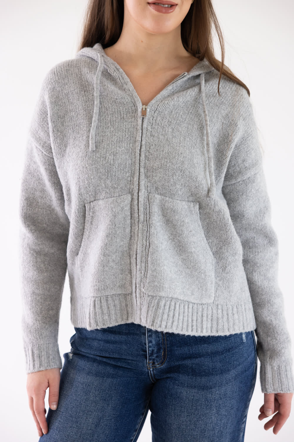 A woman wearing a light gray knit zip-up hoodie with drawstrings and front pockets, paired with blue jeans, against a plain white background.