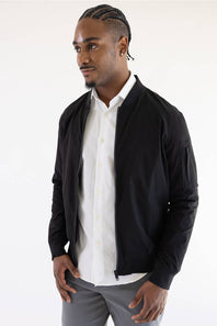 A man wearing a black bomber jacket over a white button-down shirt and gray trousers, photographed against a plain white studio background.