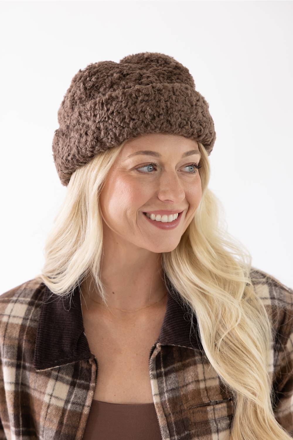 A smiling blonde woman wearing a brown chunky knit slouchy beanie and a brown plaid flannel shirt, photographed against a clean white background.