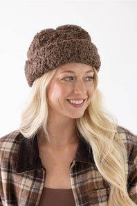 A smiling blonde woman wearing a brown chunky knit slouchy beanie and a brown plaid flannel shirt, photographed against a clean white background.