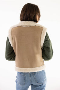 A person shown from the back wearing a shearling-lined jacket with a tan center panel, dark green sleeves, and cream-colored fleece trim, paired with blue jeans against a white background.