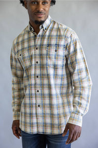 A man wearing a long-sleeve button-down plaid shirt in beige, blue, and white, paired with blue jeans, standing against a neutral gray background.