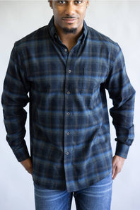A man wearing a long-sleeve button-down plaid shirt in dark blue and black, paired with blue jeans, photographed against a light gray studio background.