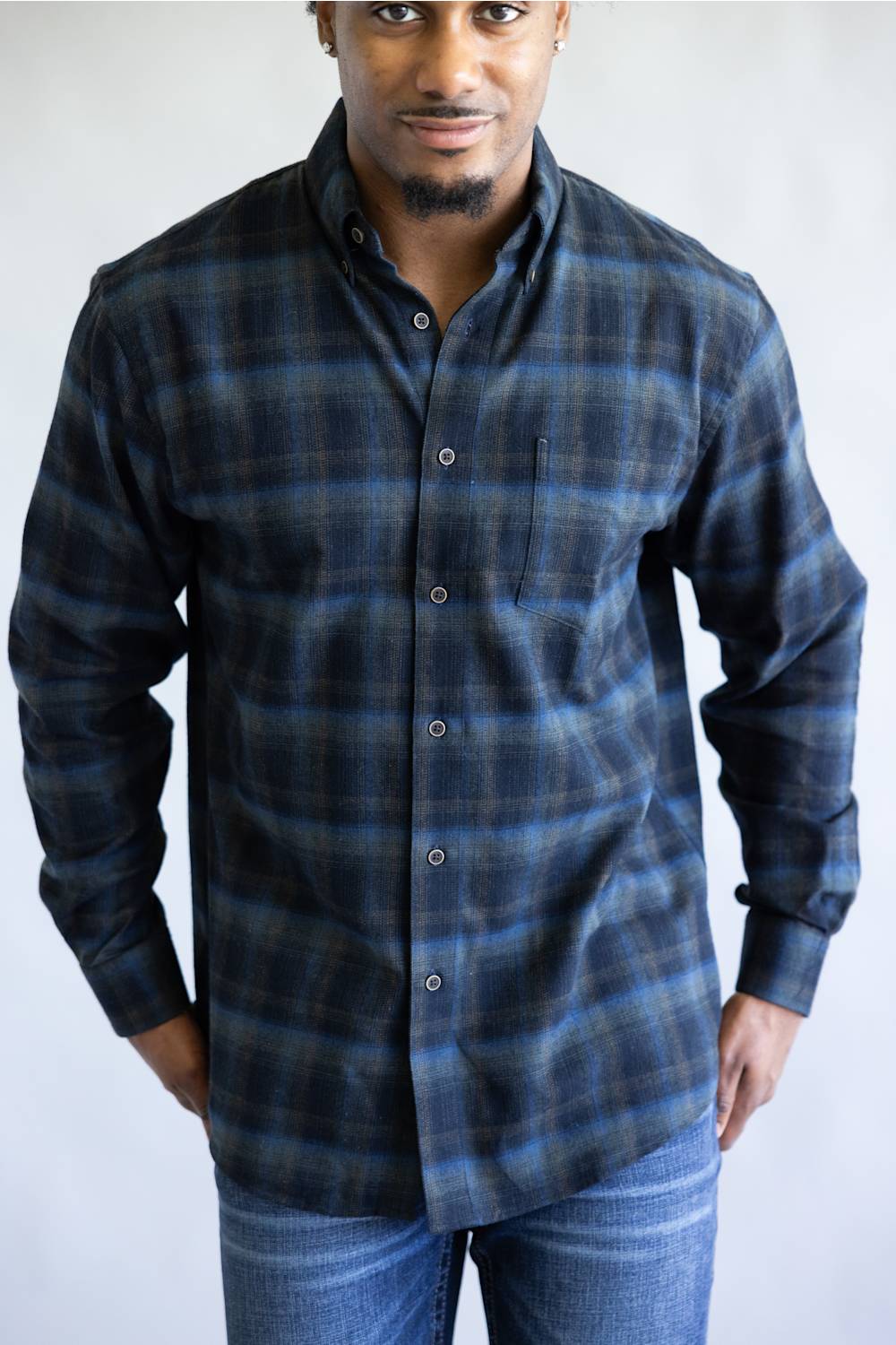 A man wearing a long-sleeve button-down plaid shirt in dark blue and black, paired with blue jeans, photographed against a light gray studio background.
