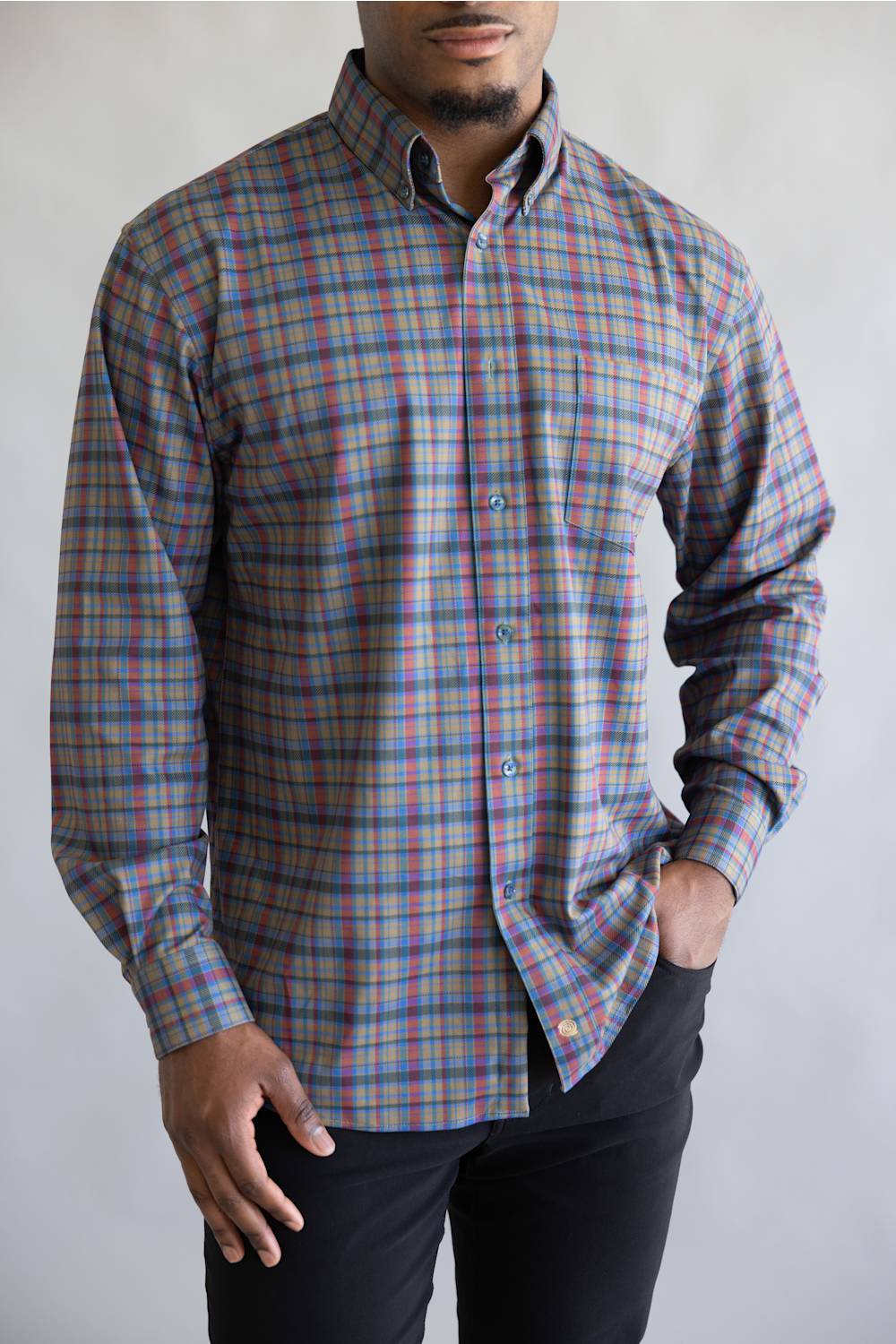 A man wearing a long-sleeve button-down plaid shirt with a multicolor pattern of blue, red, and green over a gray base, paired with black pants.