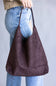 A person wearing light blue jeans and a beige top holds a large, slouchy hobo shoulder bag in dark brown faux suede against a plain gray background.