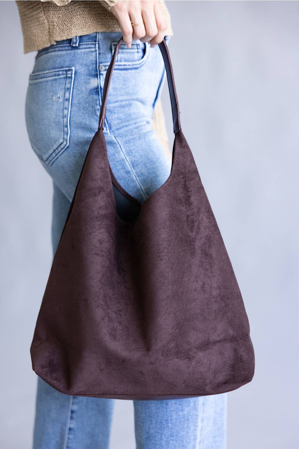 A person wearing light blue jeans and a beige top holds a large, slouchy hobo shoulder bag in dark brown faux suede against a plain gray background.