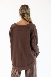 A woman viewed from behind wearing a brown long-sleeve top with raglan-style shoulder seams and striped pants, against a plain white background.