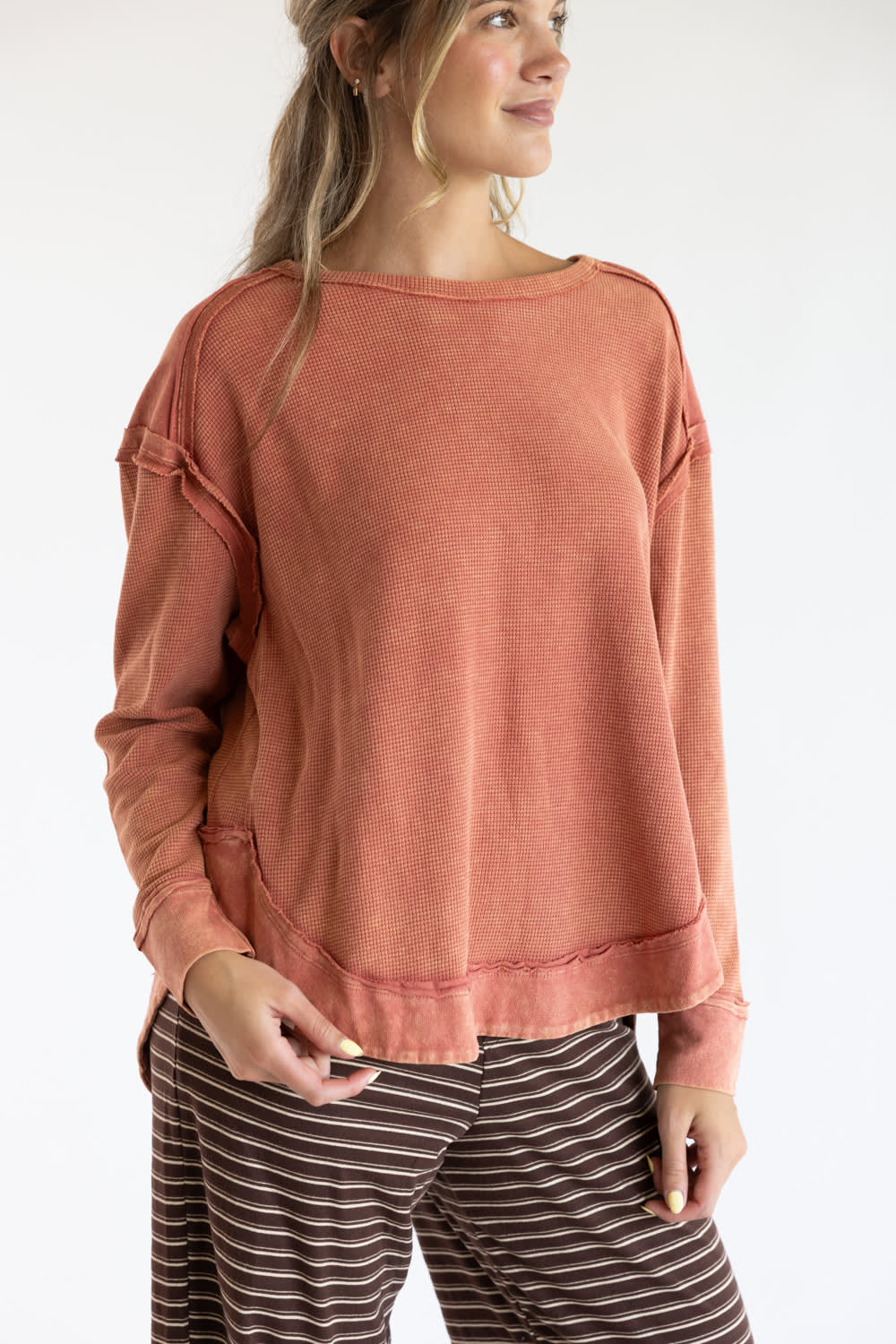 A woman wearing a long-sleeve, crew-neck top in a washed terracotta or rust color, paired with brown and white striped pants, against a plain white studio background.